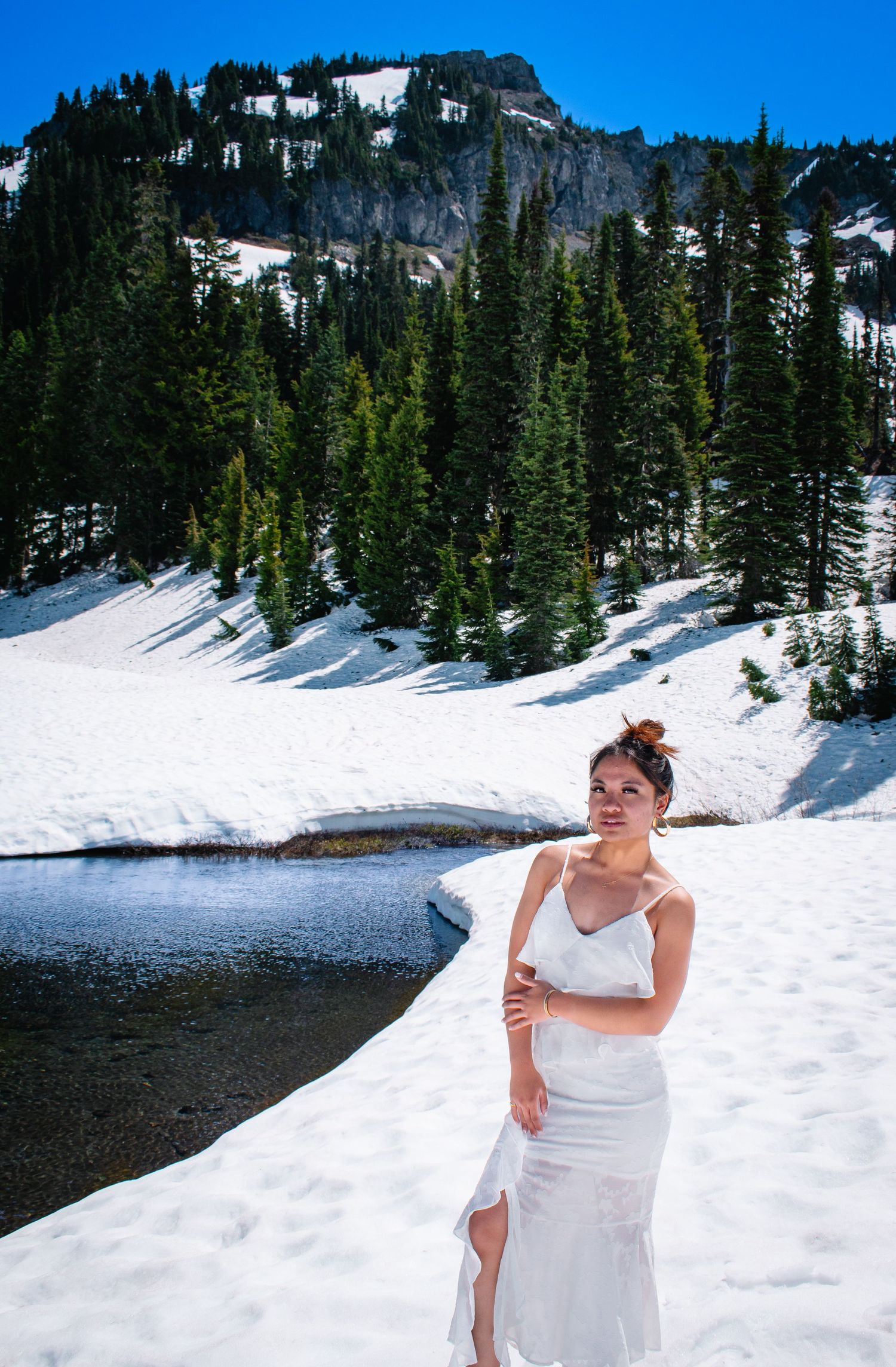 Senior portrait in a long white dress standing against the evergreen trees and snowy mountains of Washington State.