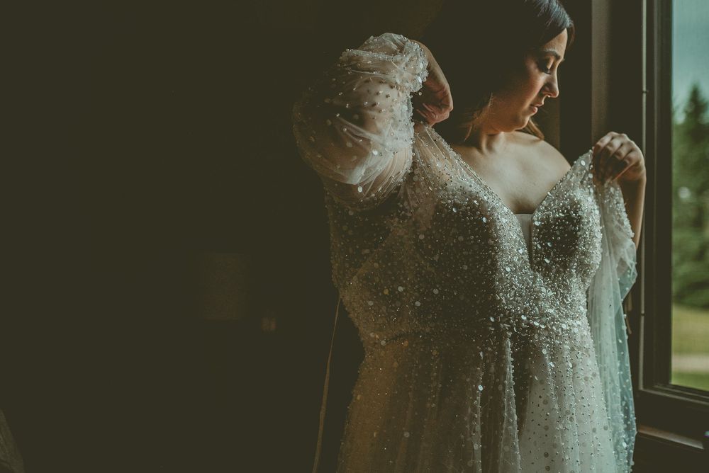 Bride in a sequined white wedding dress poses near large windows, creating a sparkly, ethereal effect. Saskatchewan Wedding Photographer