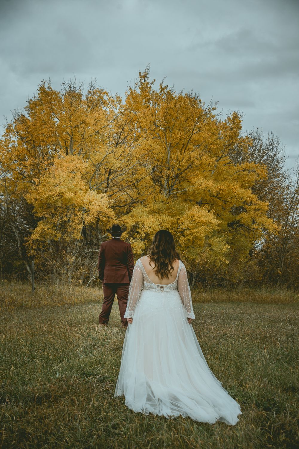 A couple in wedding attire stands in a field with golden autumn trees and moody sky in the background. Saskatchewan Wedding Photographer