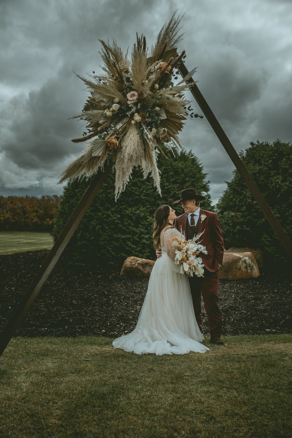 Wedding couple poses under dramatic stormy sky with dried palm leaf decorations in outdoor rustic setting. Saskatchewan Wedding Photographer