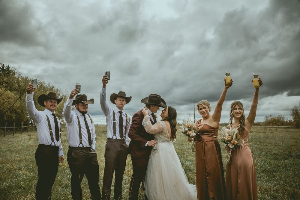 Wedding party in formal attire poses joyfully against dramatic cloudy sky during outdoor celebration. Saskatchewan Wedding Photographer