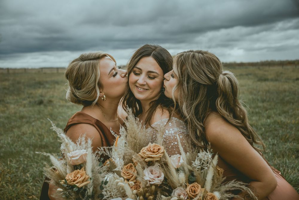 Friends share joyful moments together while holding bouquets of flowers in a serene field Saskatchewan Wedding Photographer