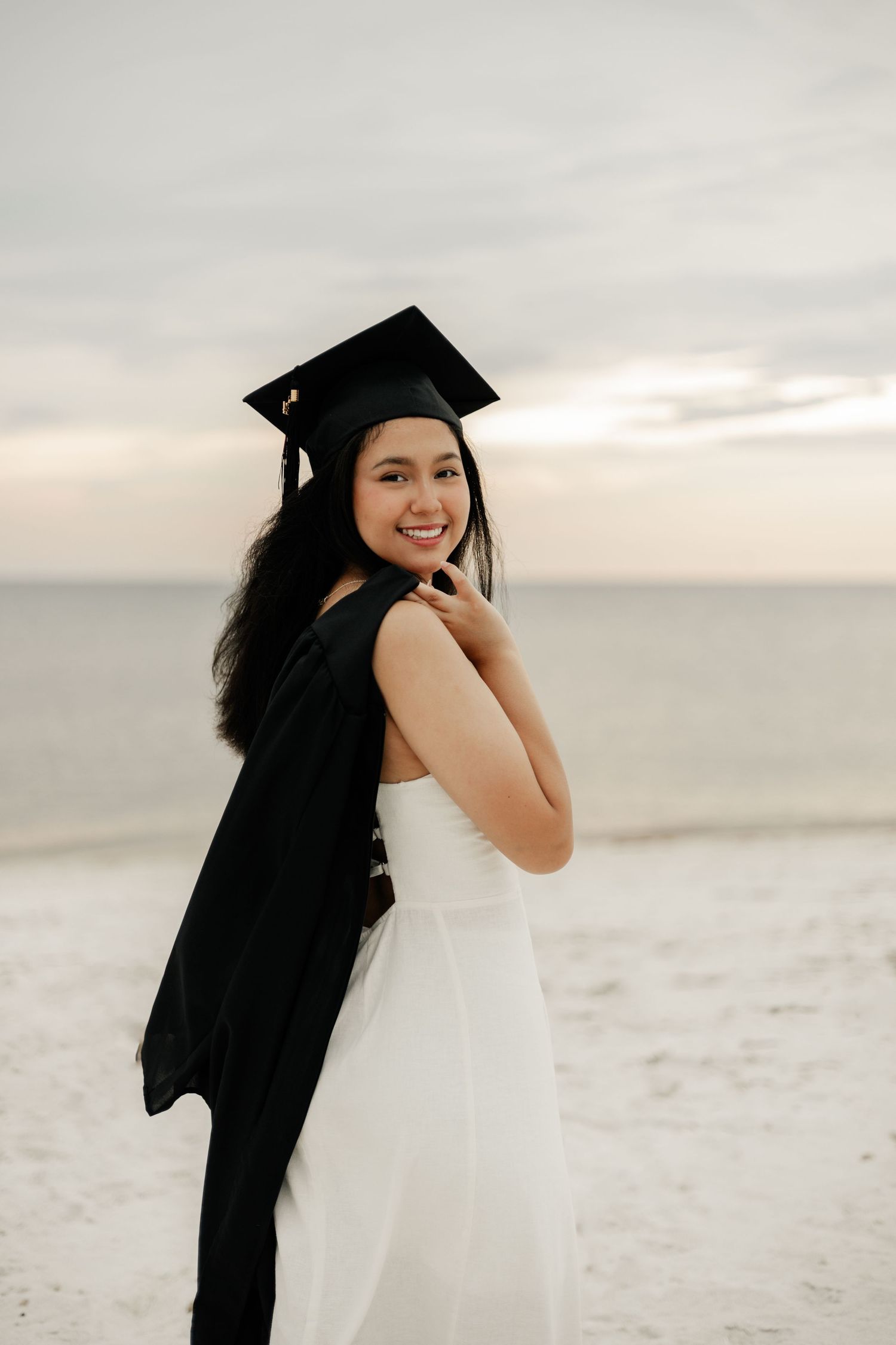 A graduate in white dress and cap poses joyfully on a sandy beach during sunset.