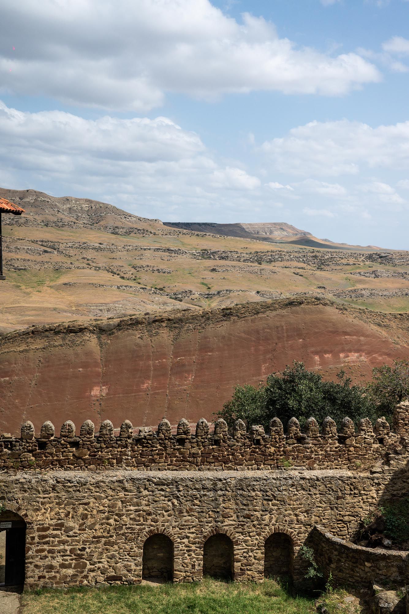 Ancient stone fortress wall with arched windows overlooks red clay hills and rugged landscape under cloudy blue sky.