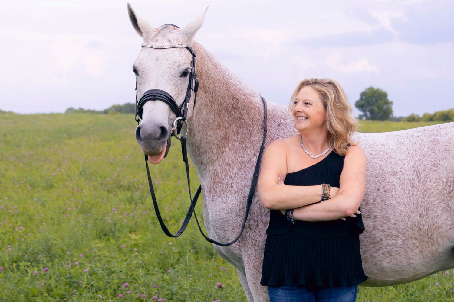 Woman in black top posing with a dappled grey horse in a sunny field.