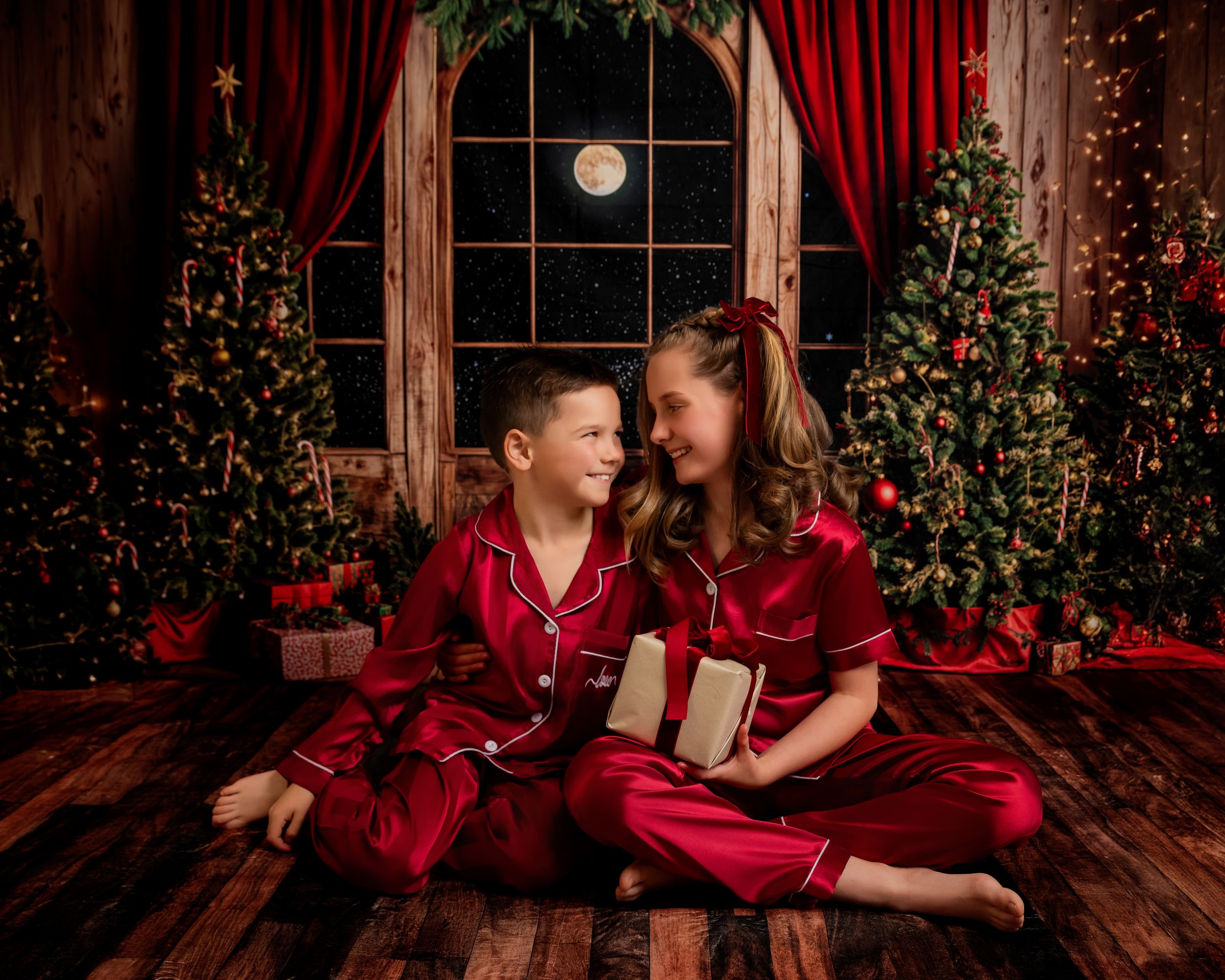 Family in matching red pajamas lounges by decorated Christmas trees during holiday photoshoot.