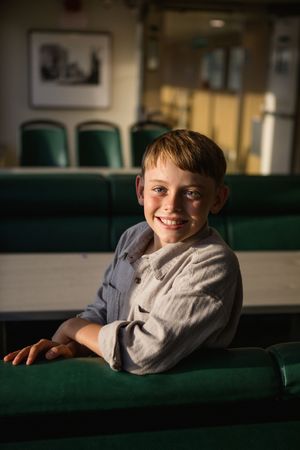 A young student sits in a green theater seat, smiling warmly in natural lighting.
