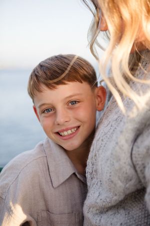 A young child smiles brightly while wearing a gray collared shirt in an outdoor portrait setting.