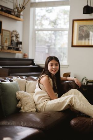 Person relaxing on a brown leather couch in a sunlit room with framed artwork and shelving visible in background.