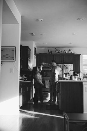 Black and white photo of two people standing in a modern kitchen with dark cabinets and sunlight streaming through the window.