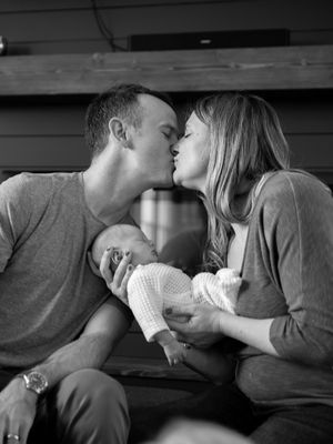 A couple shares a tender kiss while cradling their newborn baby in a black and white photograph.
