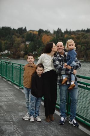 A family stands together on a bridge railing with autumn trees and water in the background on an overcast day.