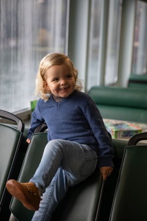 A toddler in a navy sweater and jeans sits on green seats with a playful expression.