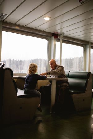 Passengers sit by large windows in a dimly lit ferry cabin during an overcast journey.