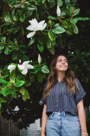 A woman wearing a striped top and denim jeans poses playfully under lush green foliage and white flowers in a garden setting.