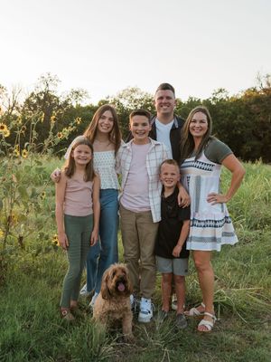 A group stands together in a grassy field at sunset with a brown dog, creating a warm family portrait moment.