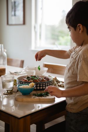 A child helps prepare food at a kitchen table with a bowl of greens and a patterned serving dish.