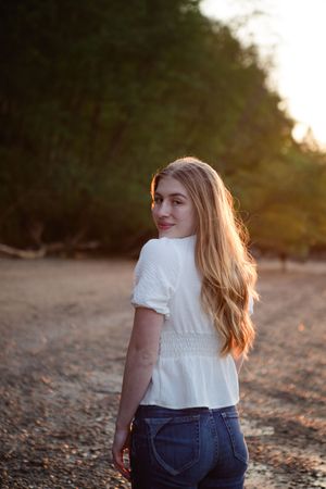 Person in white t-shirt and jeans sits on rocky beach during golden hour sunset, photographed from behind.