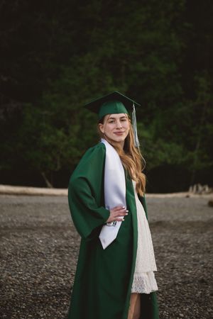 Graduate in green cap and gown standing outside in front of trees wearing a white dress for graduation photos.