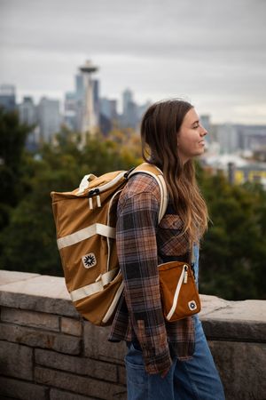 Person wearing plaid shirt and jeans with brown backpack stands against cityscape view from stone wall overlook.