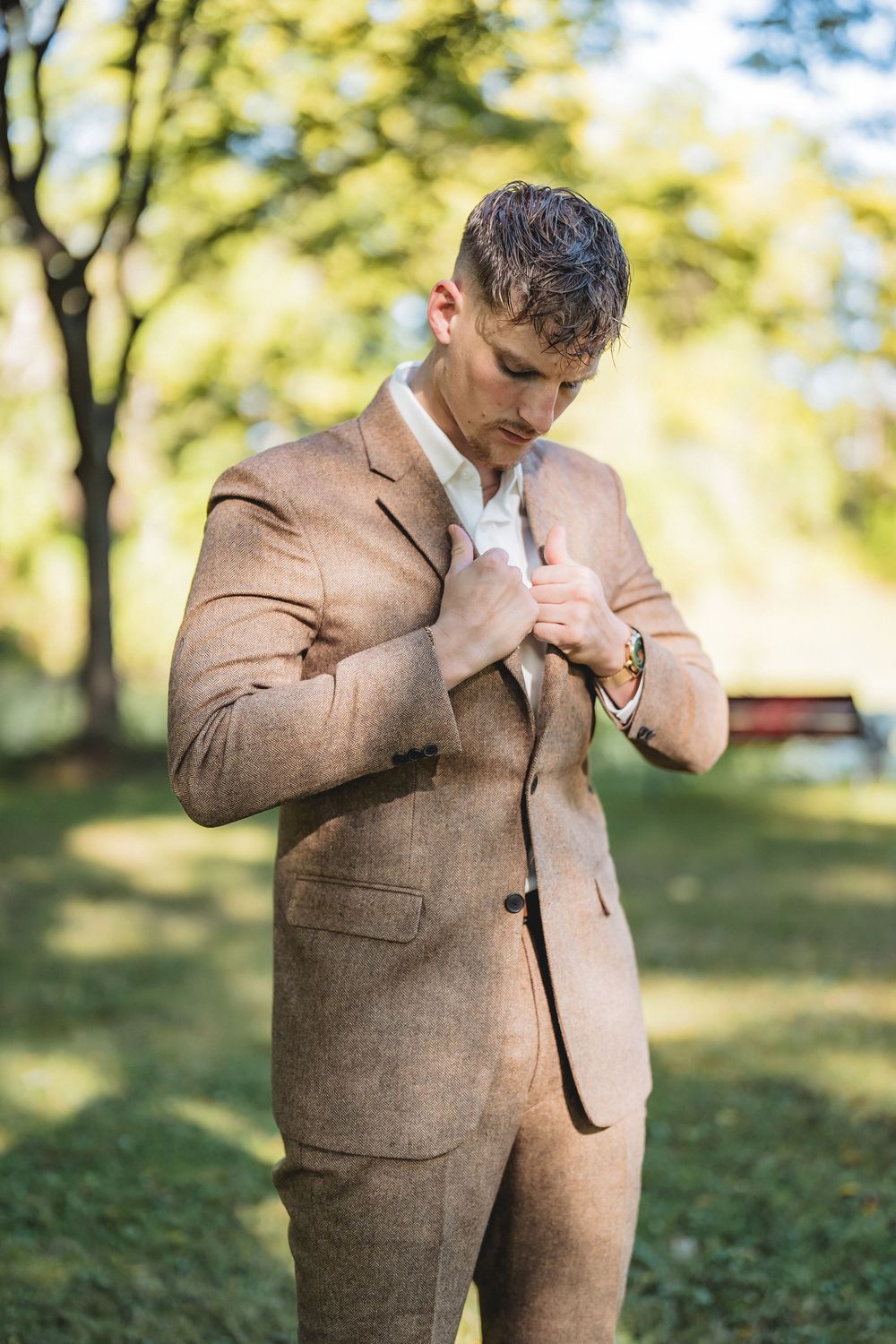 Person in brown tweed suit adjusts their jacket while standing outdoors in a park setting with trees in background.