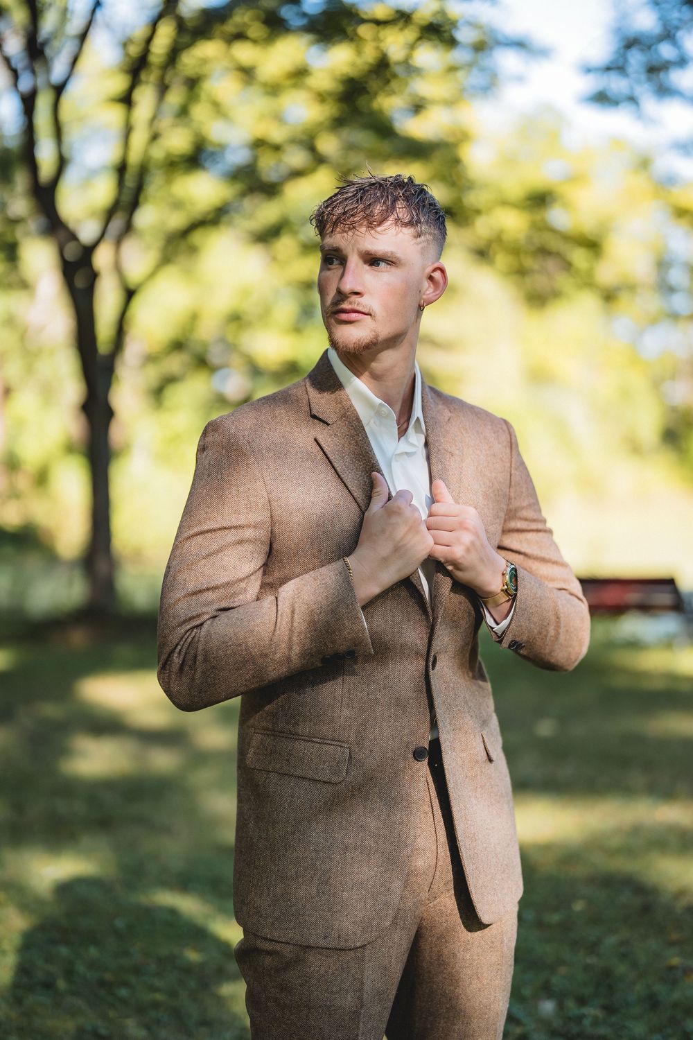 Man in brown tweed suit jacket poses against blurred autumn park background with soft natural lighting.
