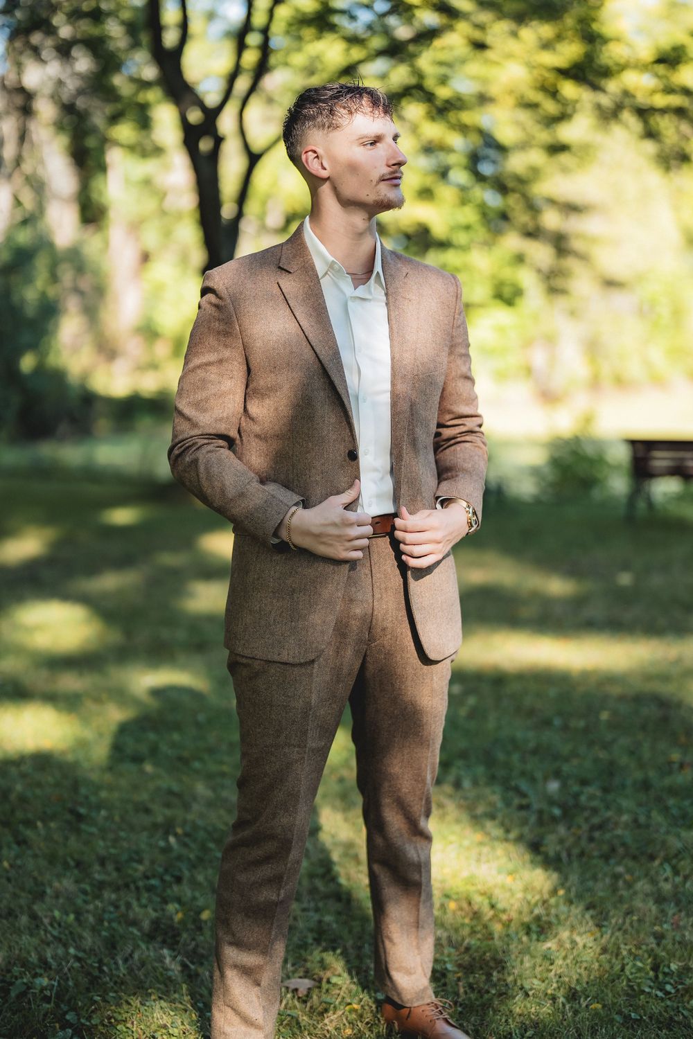 A professional wearing a brown tailored suit and white shirt poses outdoors in a sunlit garden setting.