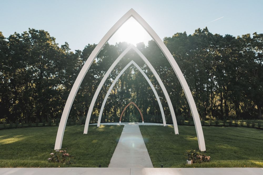 Modern white arched wedding ceremony structure stands elegantly against a lush green lawn and tree backdrop at sunset.