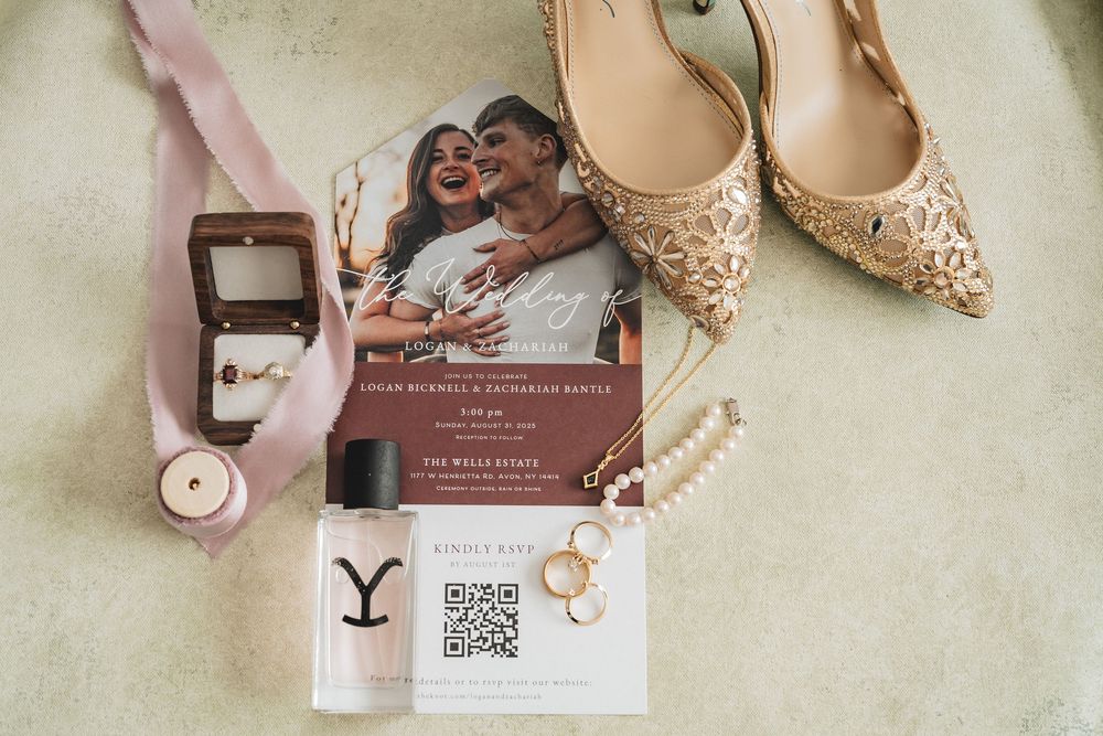 Elegant bridal accessories including blush pink heels, delicate jewelry, and wedding invitations laid on white surface.