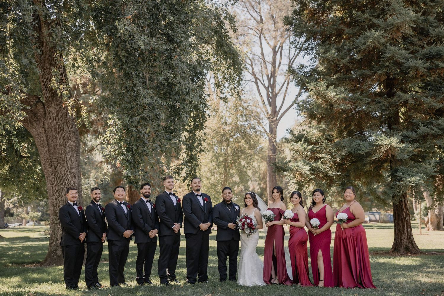 A panoramic wedding party photo in a park setting with bridesmaids in burgundy dresses and groomsmen in dark suits for Sacramento wedding.