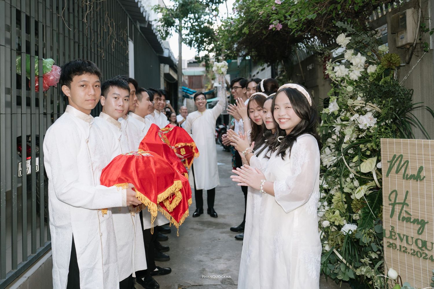 Vietnamese wedding party in white ao dai exchanges red gift boxes at flower-decorated entrance gate.