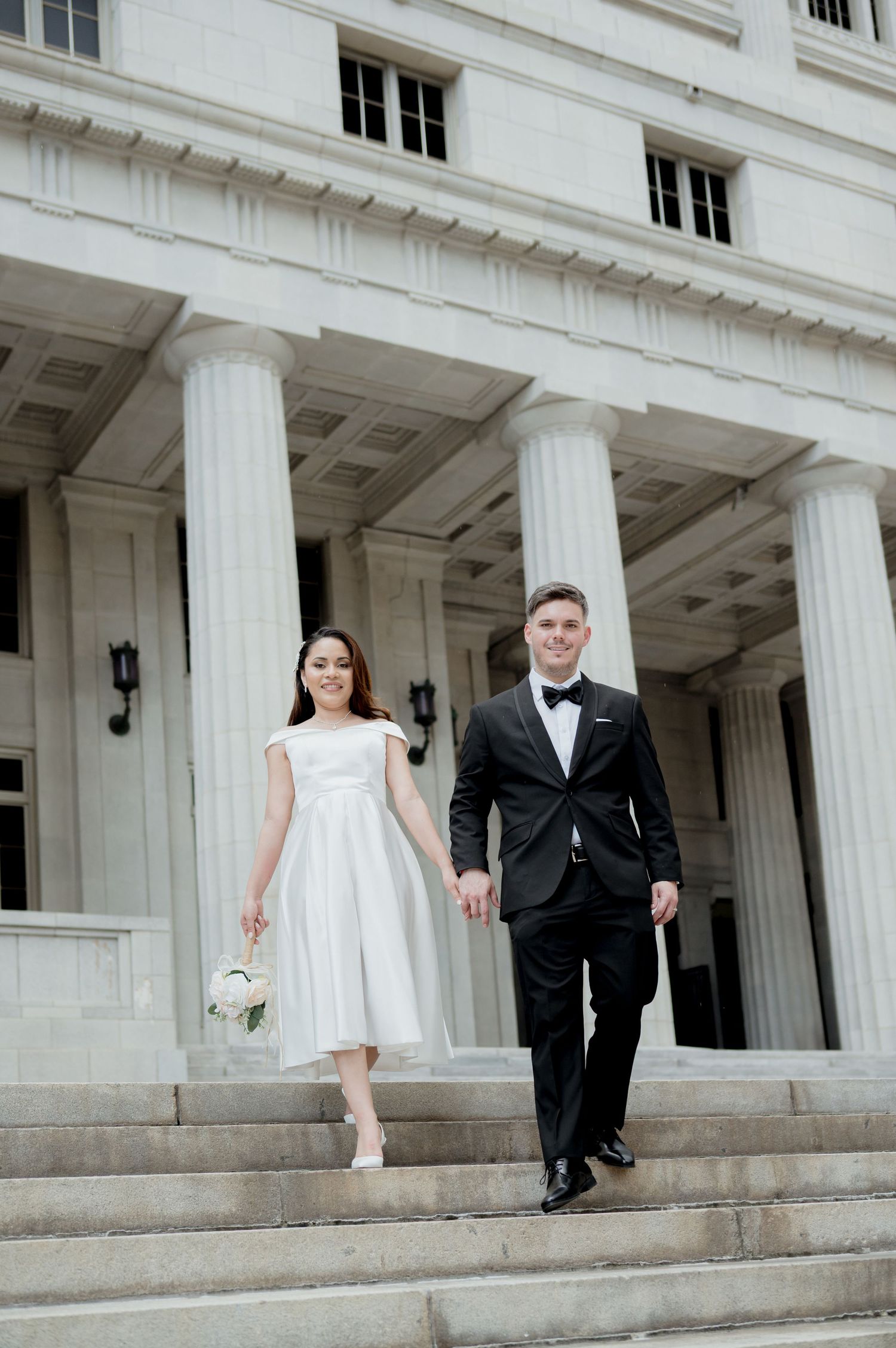 A wedding couple descends grand white stone columns and stairs of a neoclassical building in formal attire.