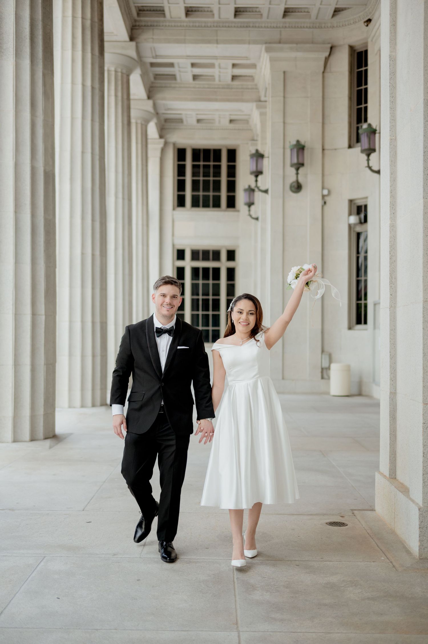 A couple in formal wedding attire playfully walks and dances through an elegant white columned hallway.