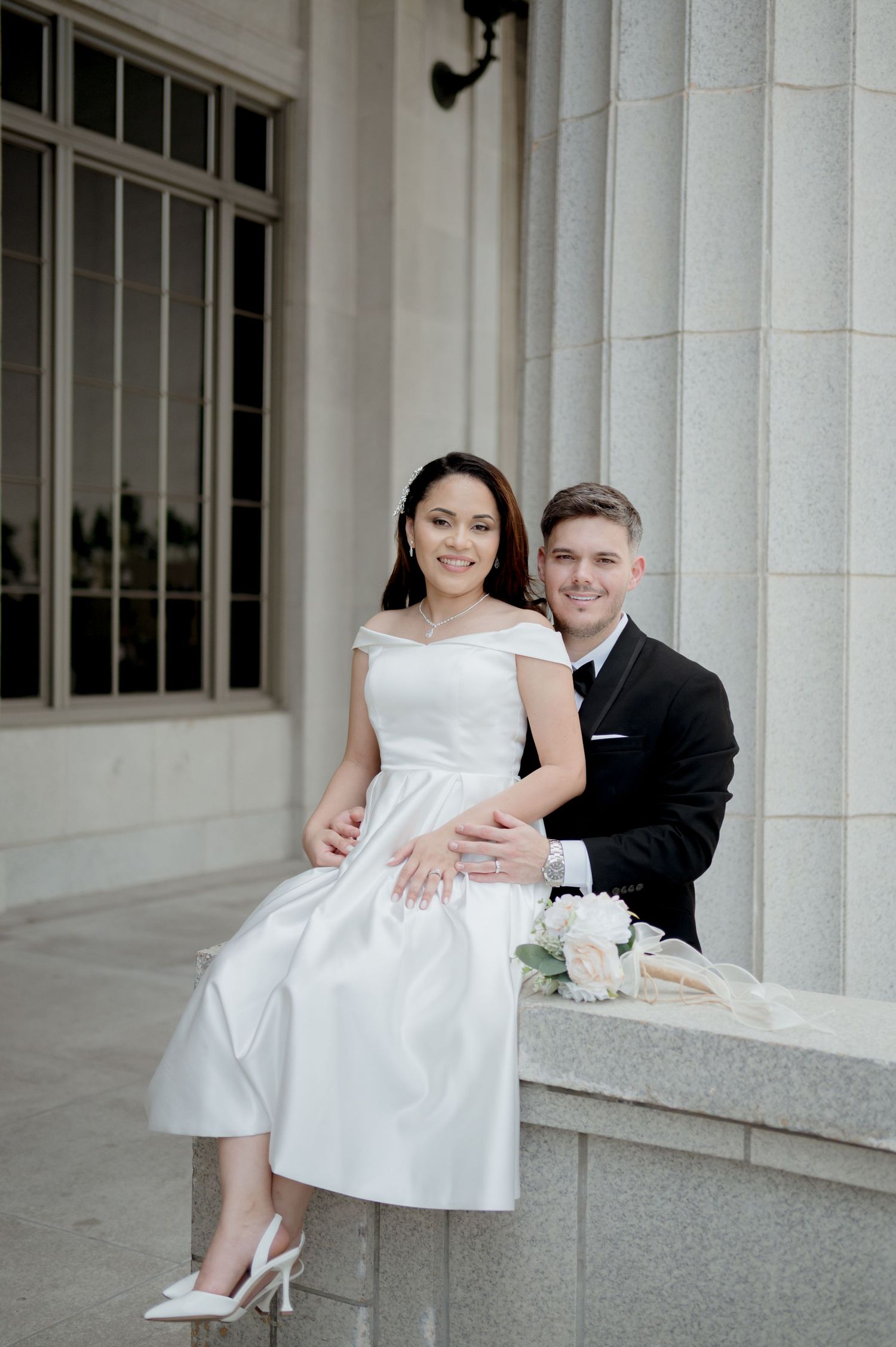 A couple in wedding attire sits together on stone steps outside a columned building during their photo session.