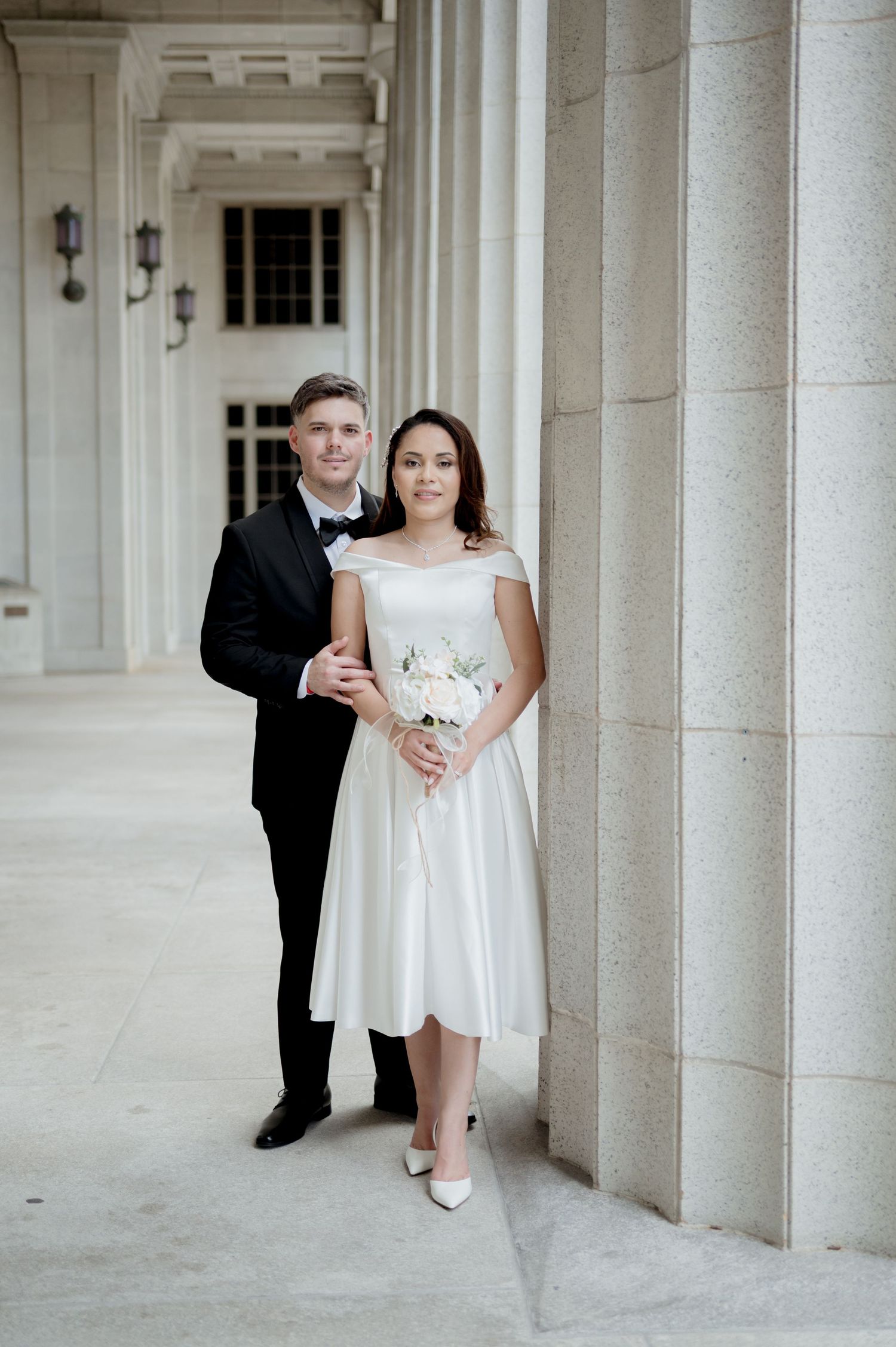 Wedding couple poses together in an elegant white colonnade hallway wearing formal attire with white flowers.