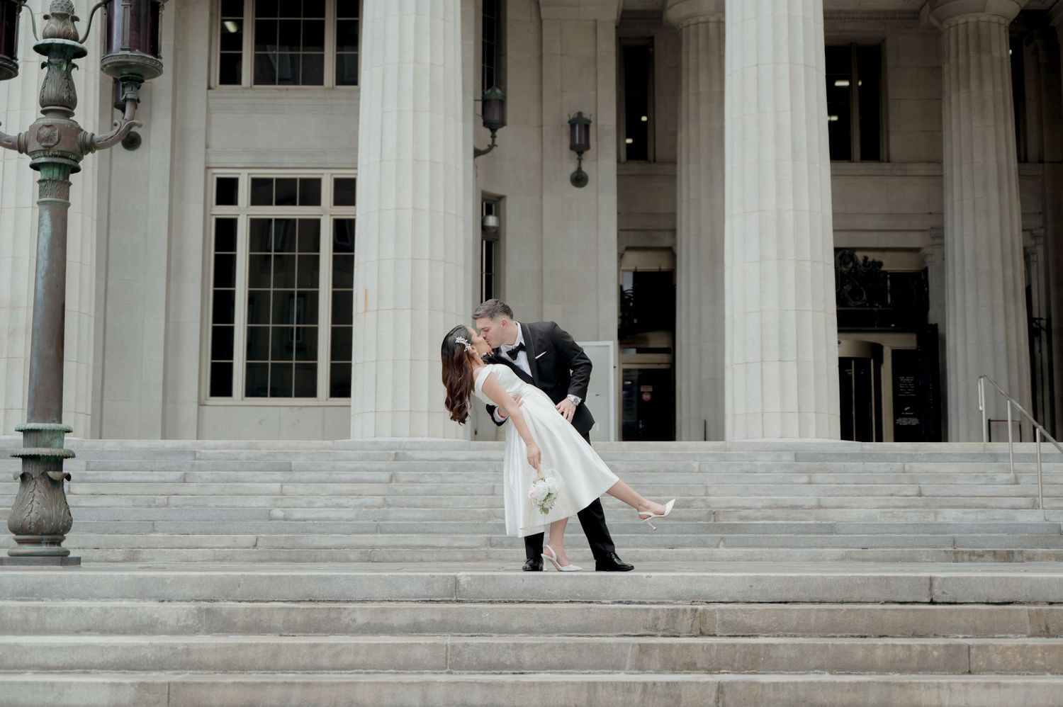 A couple shares a romantic dip while embracing in front of a grand white pillared building entrance.