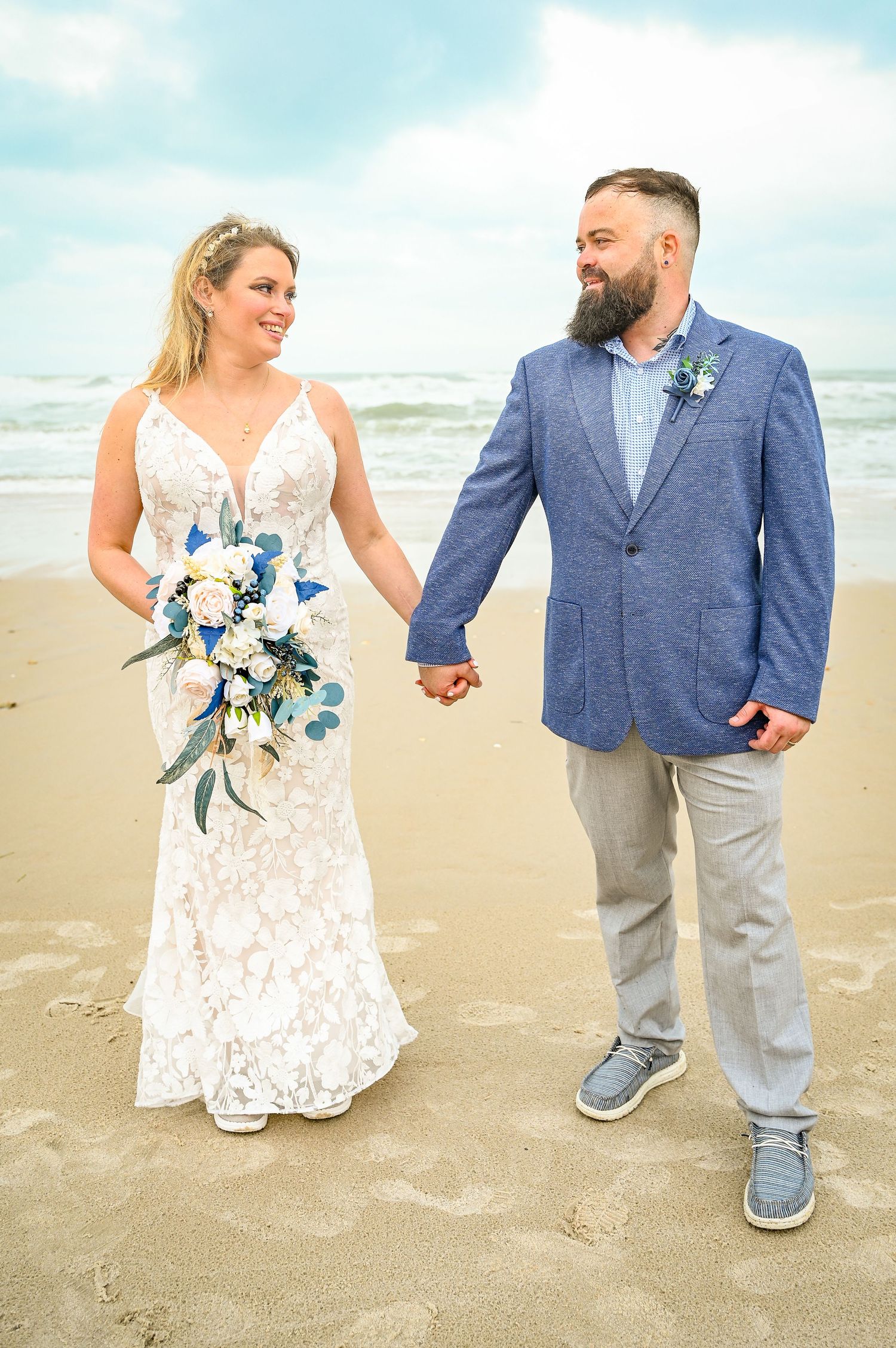 A beach wedding couple holds hands while standing on the sand, bride holding blue and white bouquet.
