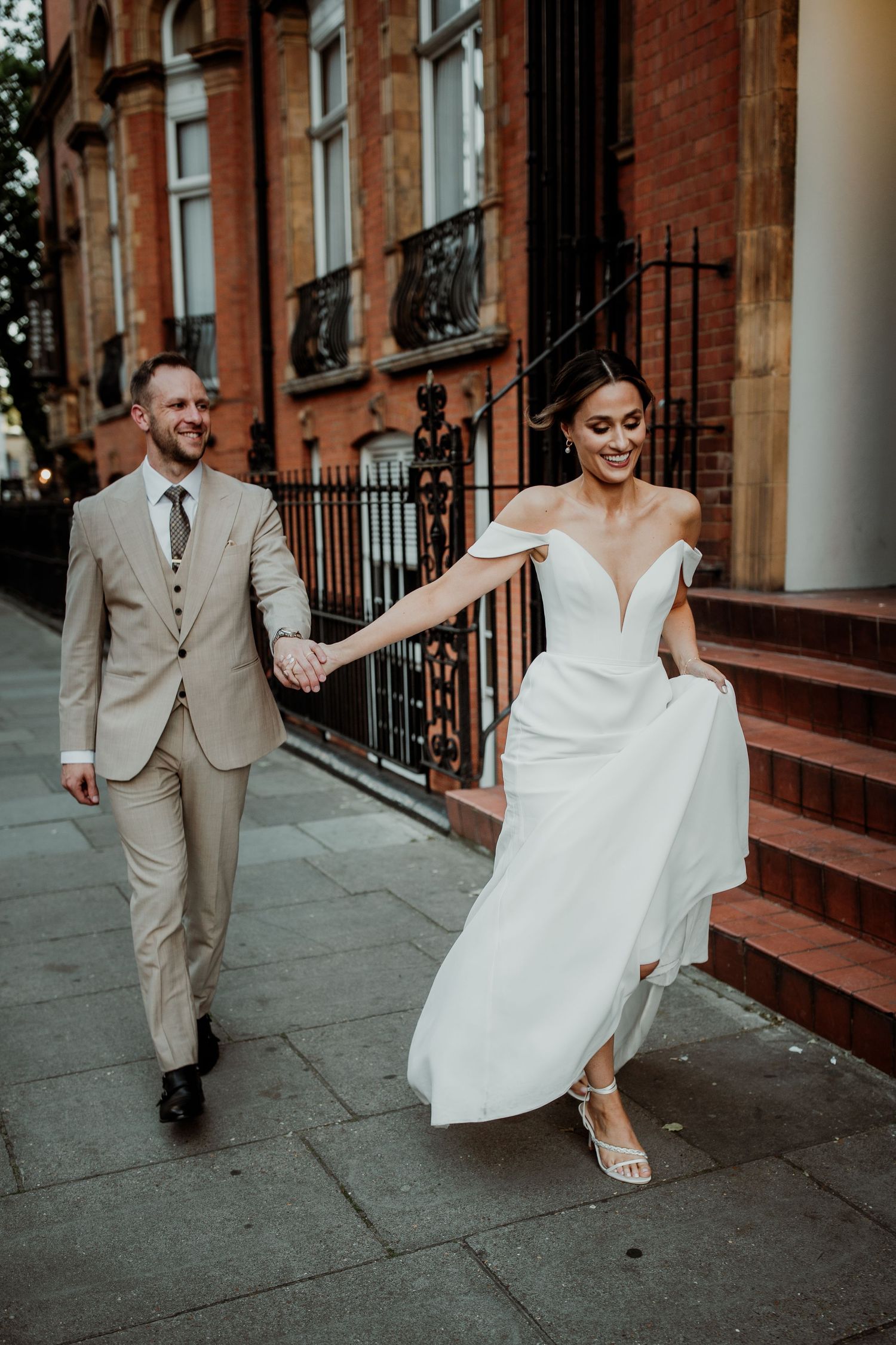 A joyful couple walks hand in hand outside a historic brick building, with the bride in a flowing white gown.
