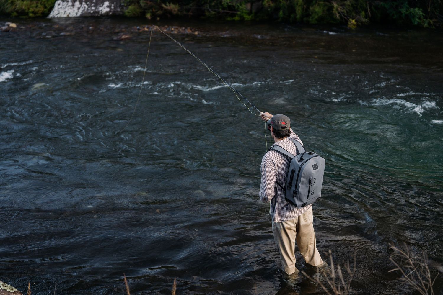 An angler stands in rushing river waters while fly fishing from the shoreline.