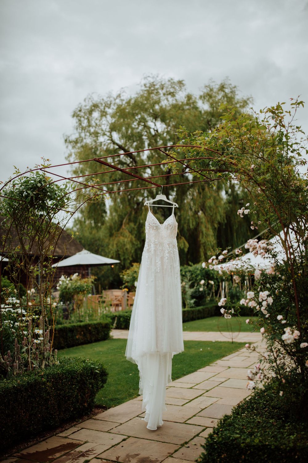 Elegant white wedding dress hanging in a garden with blooming flowers and stone pathway.