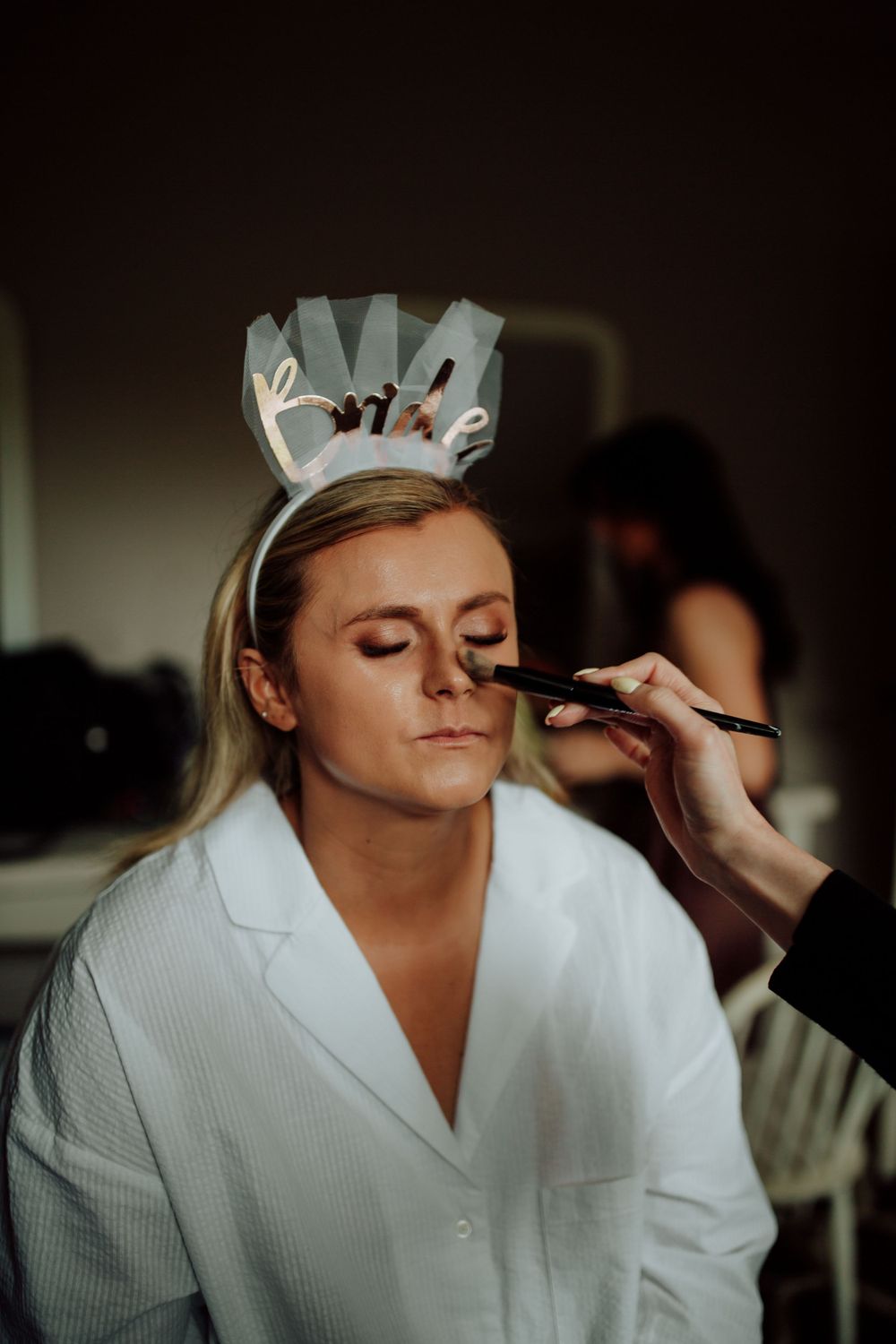 Wedding preparation scene with makeup being applied to person wearing white robe and crown.
