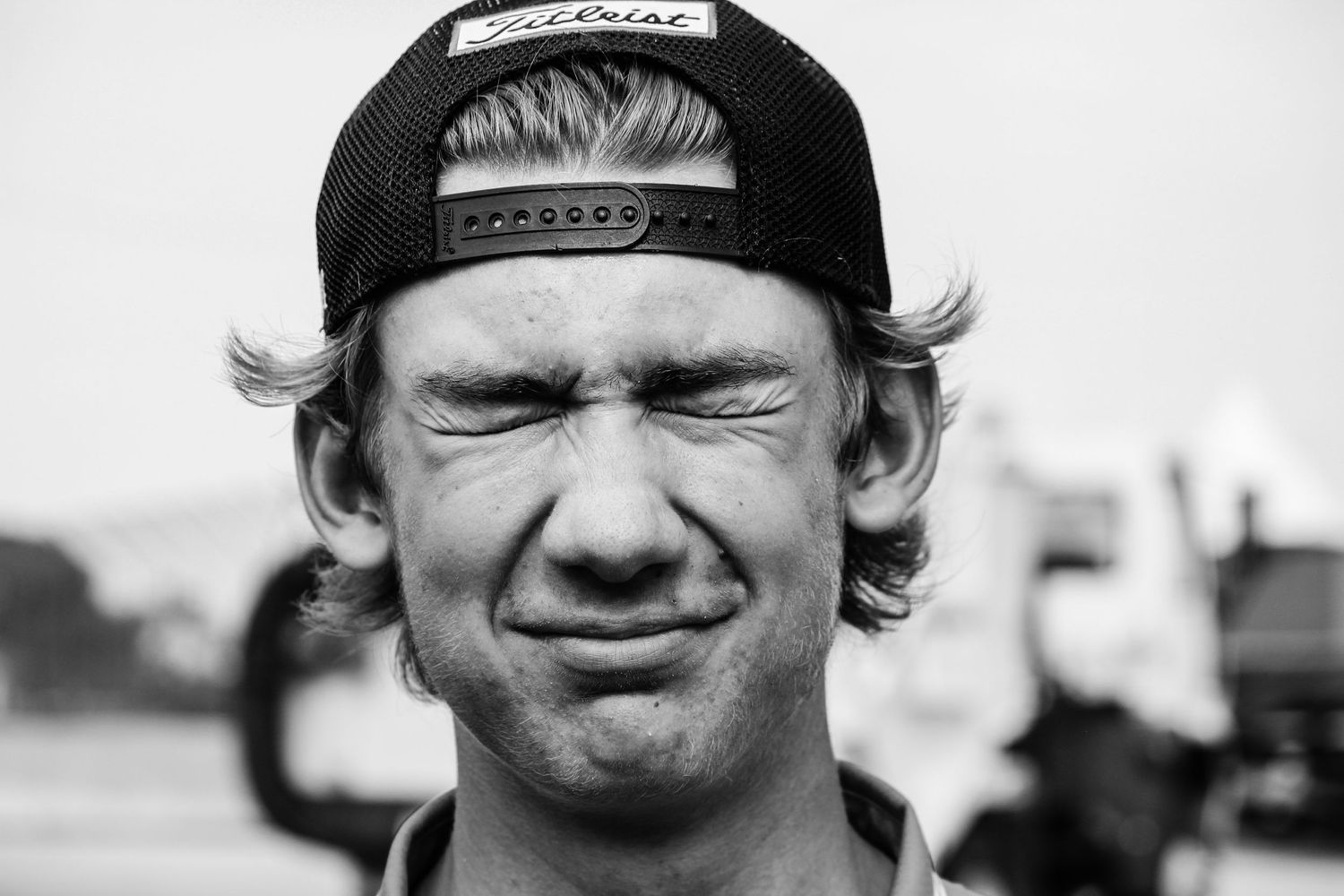 Black and white image of a young man wearing a backwards hat with his eyes closed and a playful grin on his face.