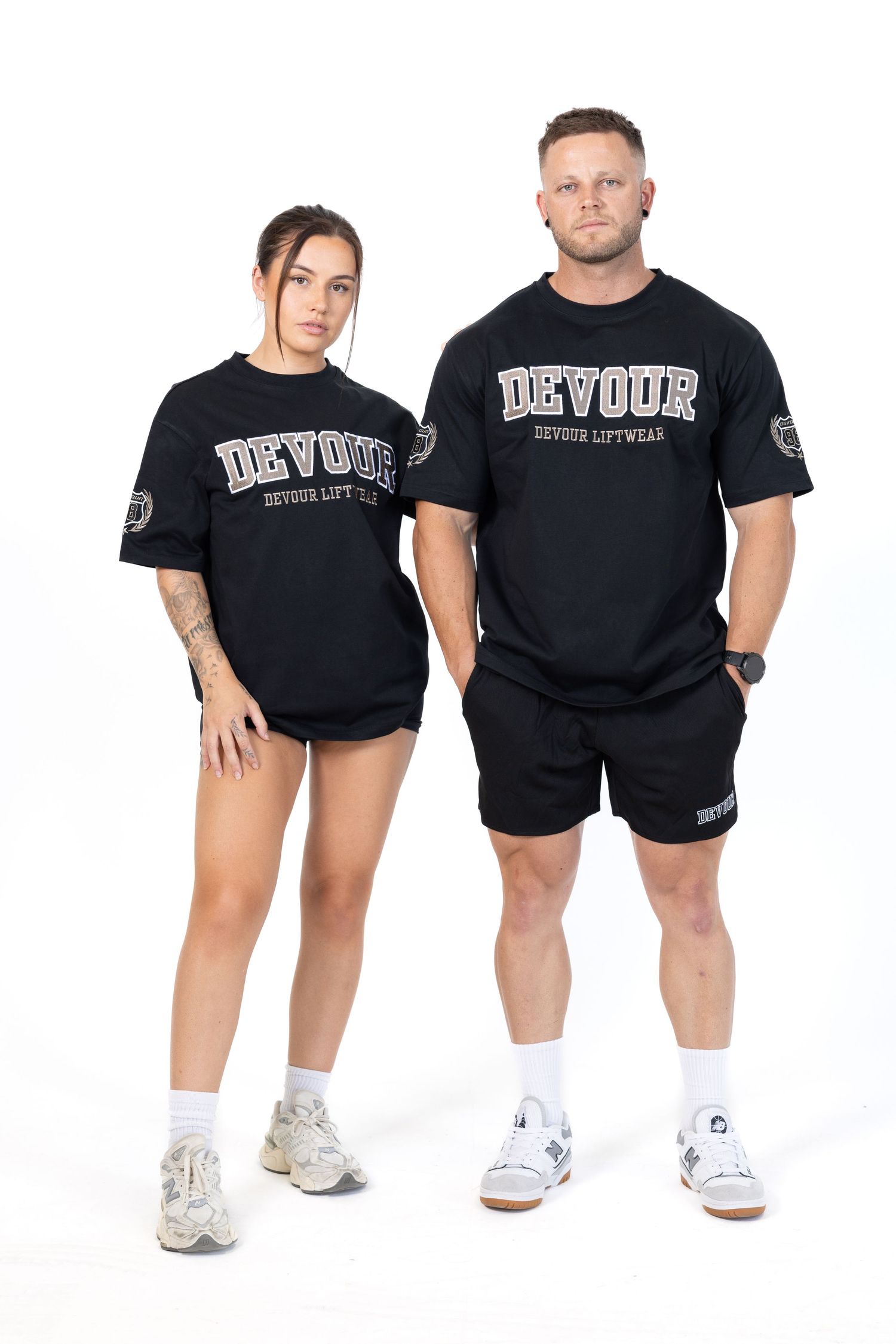 Two tattooed men in black clothing posing in a studio setting against a white background.