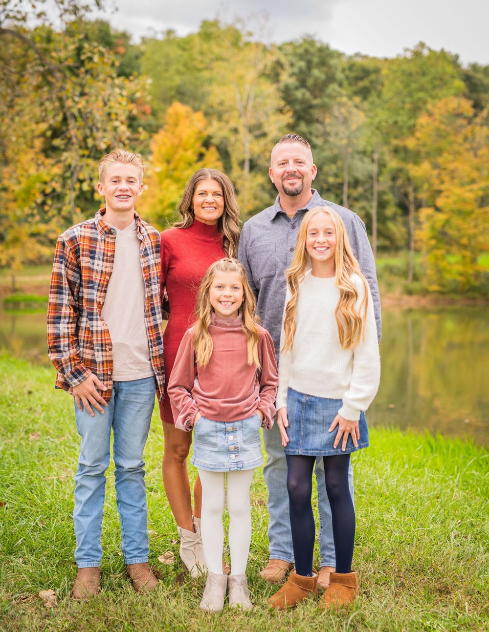 Family group poses together in casual clothing against autumn trees and lake backdrop.