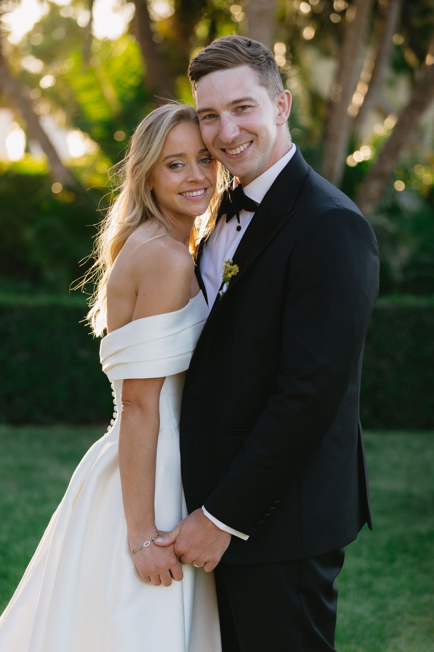 A bride in an off-shoulder white gown and groom in black tuxedo embrace outdoors with golden sunlight filtering through trees.