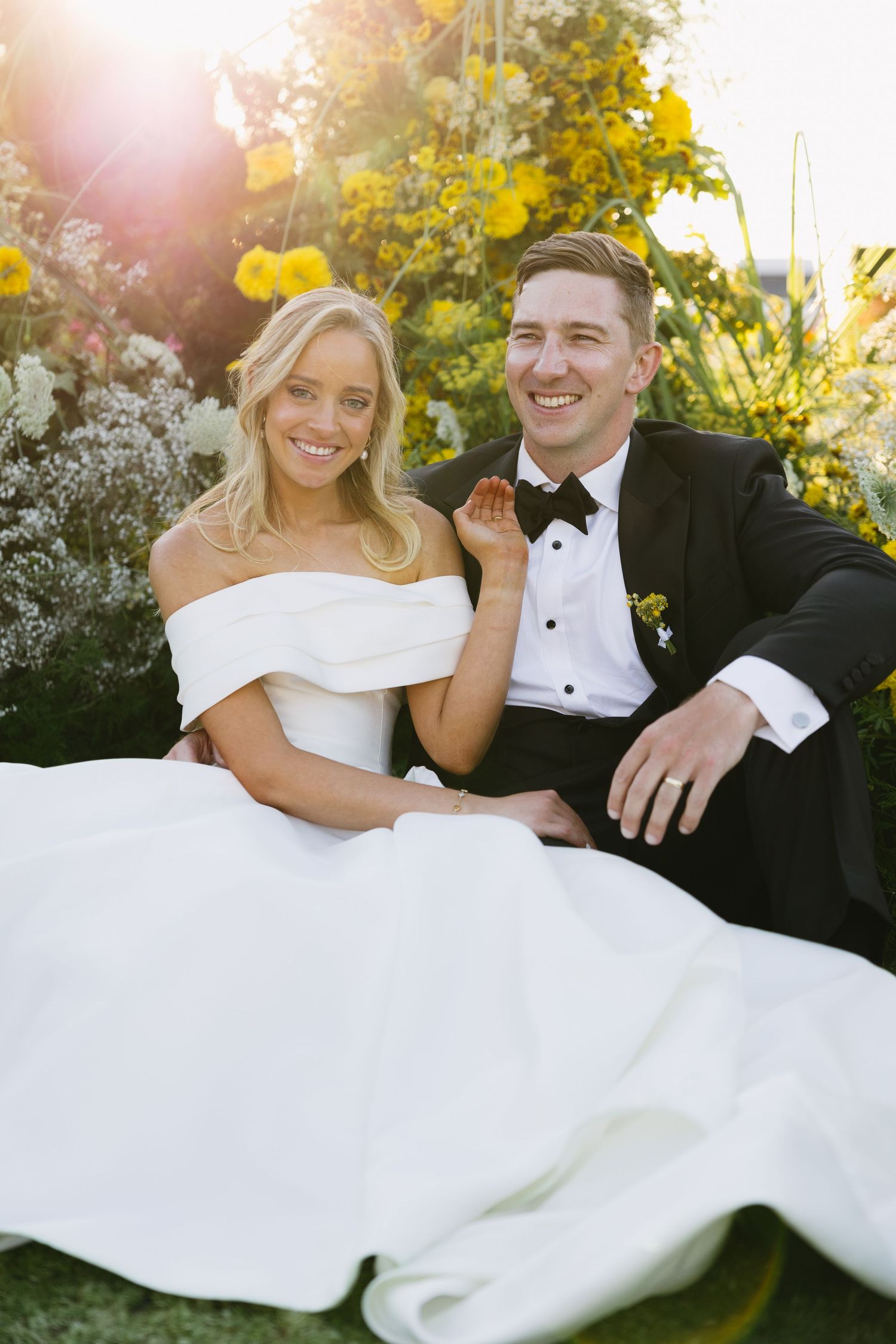 A wedding couple sits together on grass during golden hour with yellow flowers and sunlight in the background.