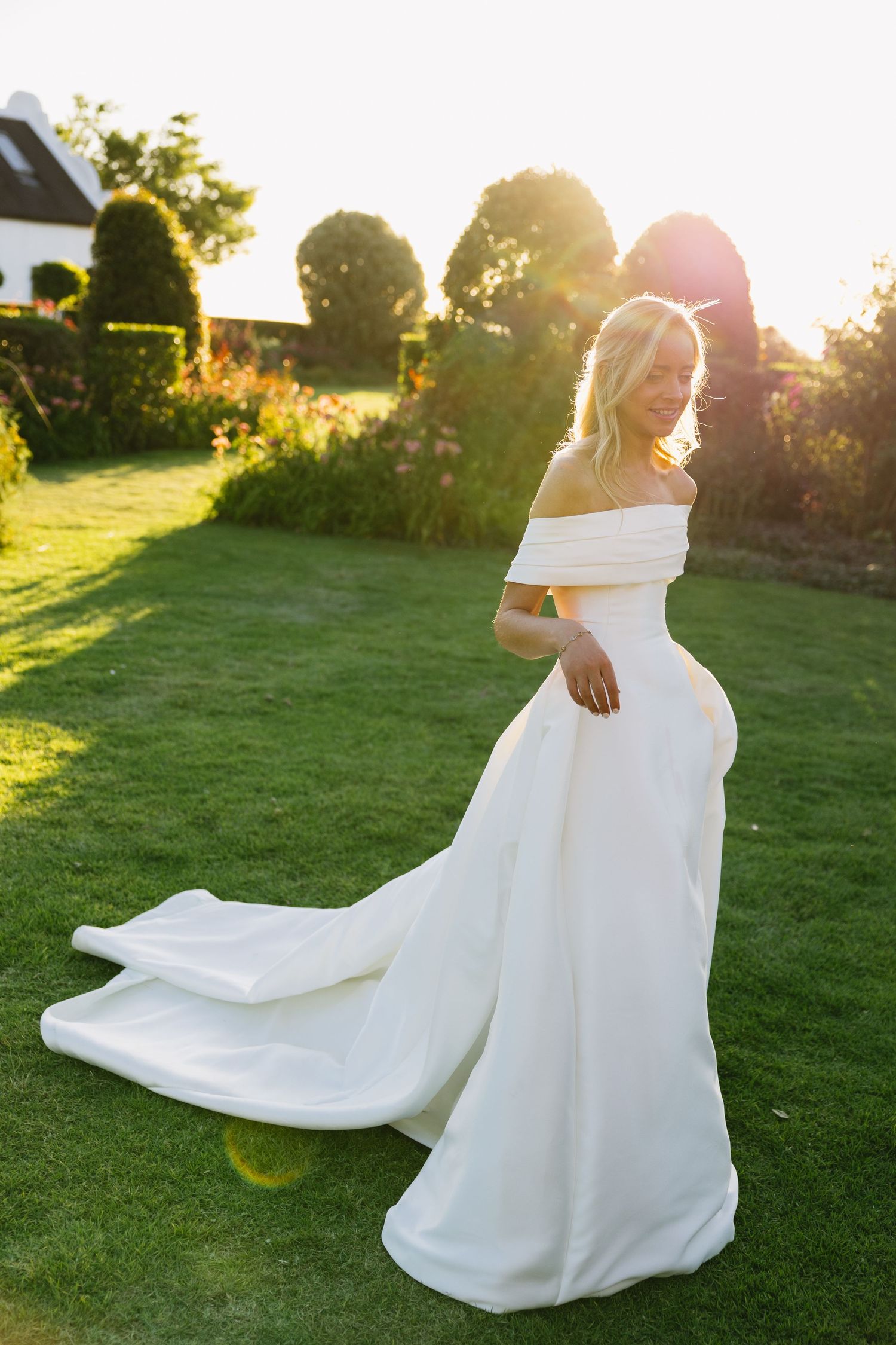 An elegant off-shoulder wedding dress flows gracefully on a lawn during sunset with garden scenery.