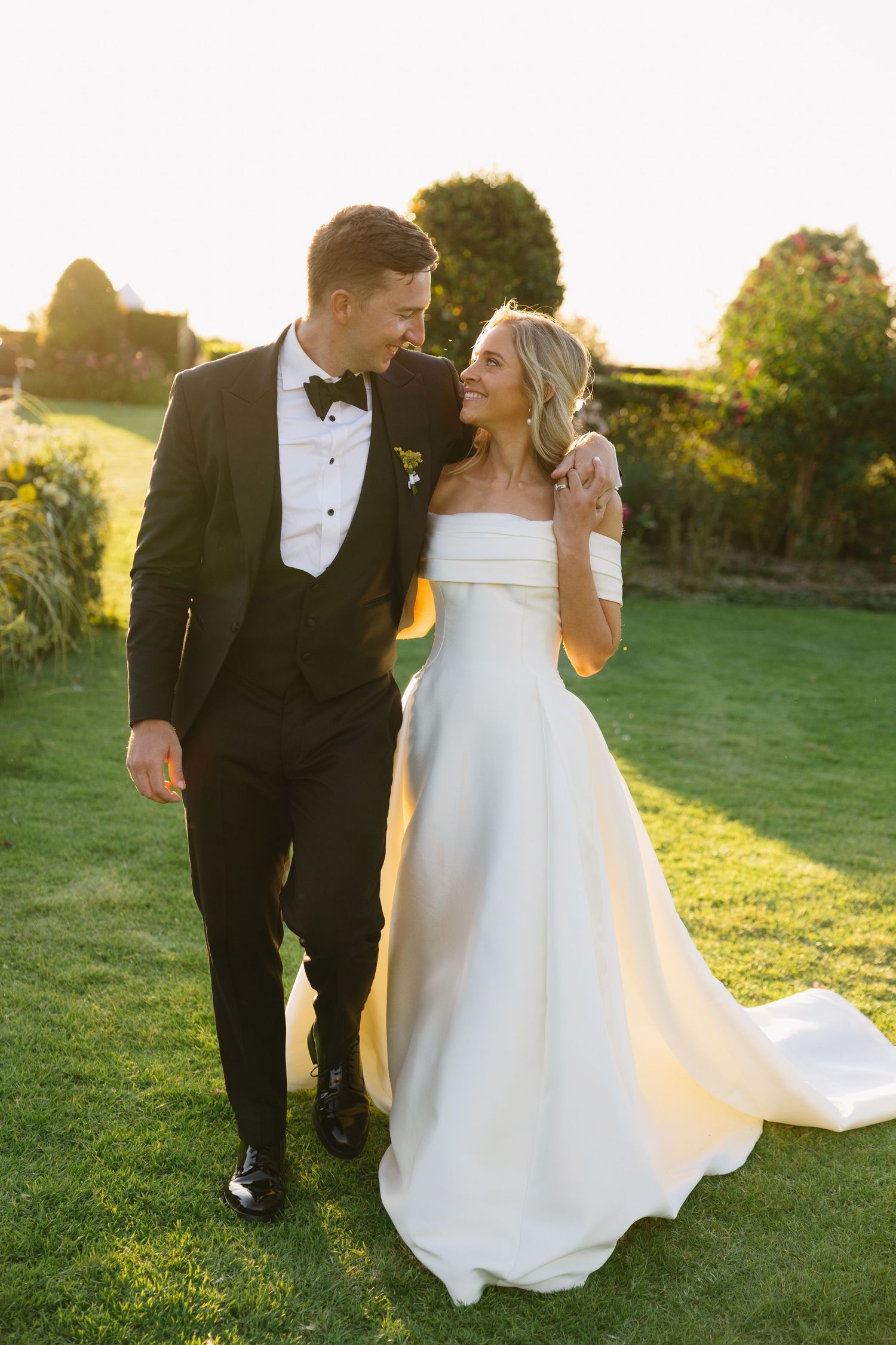 A couple in wedding attire walks together on a grassy lawn during golden hour sunset.