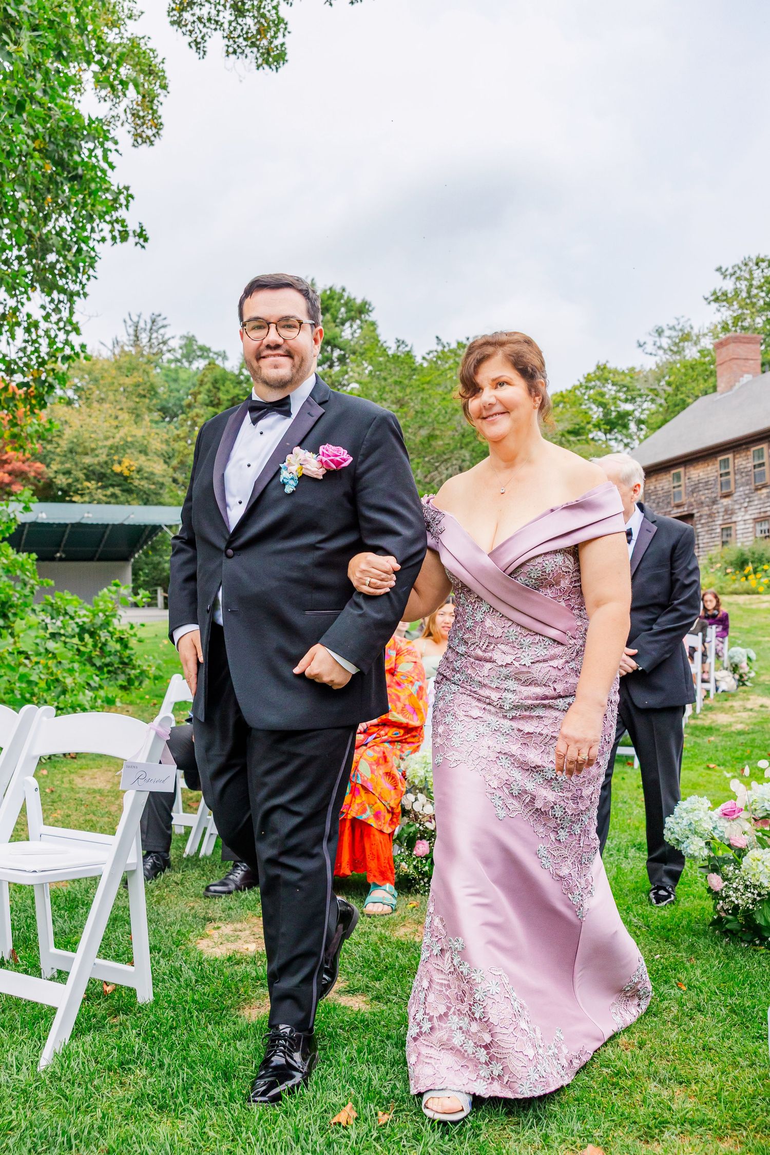 A formal outdoor wedding party poses on a grassy lawn with white chairs and trees in the background.