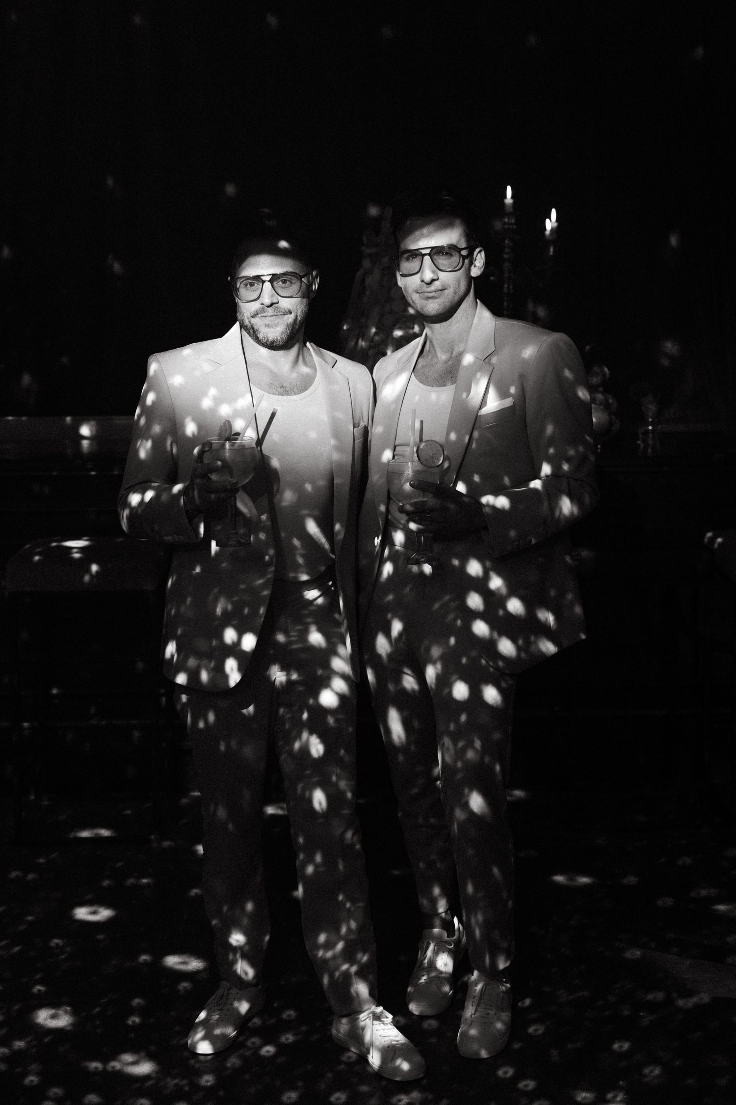 Two people in dark suits stand together while bright bokeh lights create a dreamy nighttime atmosphere.
